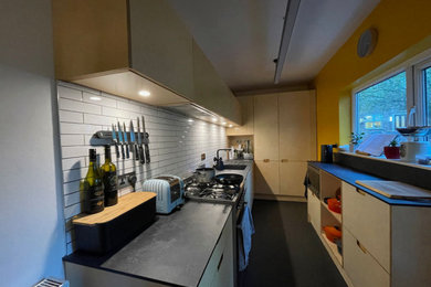 This is an example of a small midcentury grey and white galley enclosed kitchen in Manchester with a submerged sink, flat-panel cabinets, light wood cabinets, composite countertops, white splashback, cement tile splashback, stainless steel appliances, vinyl flooring, a breakfast bar, black floors, black worktops, a drop ceiling and feature lighting.