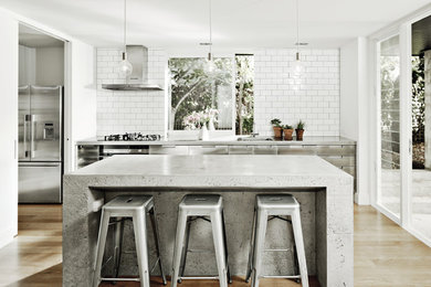 This is an example of a mid-sized industrial single-wall kitchen with flat-panel cabinets, stainless steel cabinets, concrete benchtops, white splashback, subway tile splashback, stainless steel appliances, light hardwood floors, with island, beige floor and an integrated sink.