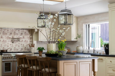 Example of a beach style medium tone wood floor, beige floor and tray ceiling kitchen design in Orange County with a farmhouse sink, beaded inset cabinets, soapstone countertops, stone tile backsplash, stainless steel appliances, an island and black countertops