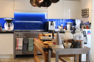 Inspiration for a large eclectic u-shaped open plan kitchen in London with a drop-in sink, flat-panel cabinets, white cabinets, granite benchtops, blue splashback, glass sheet splashback, stainless steel appliances, concrete floors and with island.