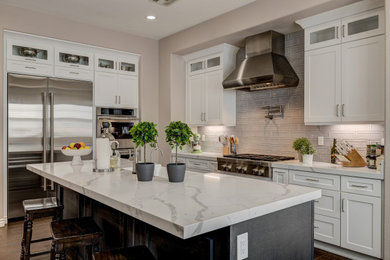 Inspiration for a transitional l-shaped dark wood floor and brown floor kitchen remodel in Las Vegas with a farmhouse sink, shaker cabinets, white cabinets, gray backsplash, stainless steel appliances, an island and white countertops