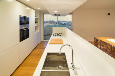 This is an example of a modern galley eat-in kitchen in Venice with a drop-in sink, flat-panel cabinets, white cabinets, quartzite benchtops, white splashback, stainless steel appliances, light hardwood floors, with island and white benchtop.