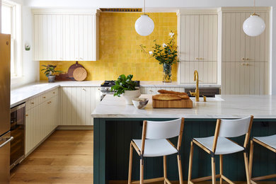 Large transitional l-shaped medium tone wood floor and brown floor eat-in kitchen photo in San Francisco with a farmhouse sink, flat-panel cabinets, beige cabinets, quartzite countertops, yellow backsplash, terra-cotta backsplash, stainless steel appliances, an island and white countertops