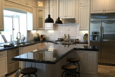 Design ideas for a mid-sized traditional l-shaped eat-in kitchen in Orange County with a double-bowl sink, raised-panel cabinets, white cabinets, granite benchtops, white splashback, ceramic splashback, stainless steel appliances, travertine floors, with island, beige floor and black benchtop.