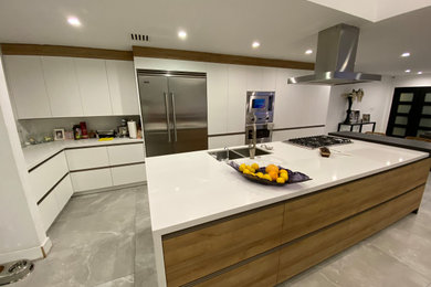 Example of a huge minimalist galley porcelain tile, gray floor and vaulted ceiling eat-in kitchen design in Los Angeles with a drop-in sink, flat-panel cabinets, white cabinets, quartzite countertops, white backsplash, stainless steel appliances and an island