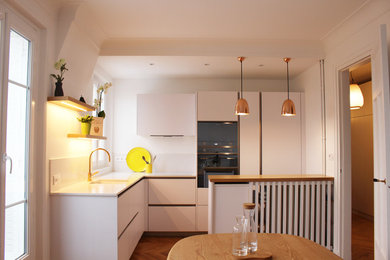 Cette photo montre une petite cuisine américaine tendance en L avec un évier encastré, un placard avec porte à panneau encastré, des portes de placard blanches, plan de travail en marbre, une crédence blanche, une crédence en marbre, un électroménager noir, parquet clair, îlot, un sol marron et un plan de travail blanc.