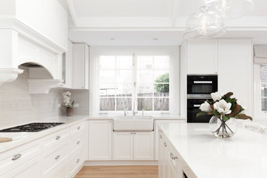 Design ideas for a mid-sized l-shaped open plan kitchen in Sydney with a farmhouse sink, shaker cabinets, white cabinets, quartz benchtops, beige splashback, ceramic splashback, black appliances, light hardwood floors, with island and white benchtop.