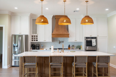 Example of a cottage single-wall medium tone wood floor and brown floor open concept kitchen design in Austin with a drop-in sink, recessed-panel cabinets, white cabinets, marble countertops, white backsplash, marble backsplash, stainless steel appliances, an island and white countertops
