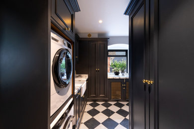 This is an example of a large classic l-shaped separated utility room in Hertfordshire with a belfast sink, shaker cabinets, black cabinets, engineered stone countertops, white splashback, engineered quartz splashback, ceramic flooring, a stacked washer and dryer, multi-coloured floors and white worktops.