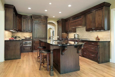 Inspiration for a large timeless u-shaped light wood floor and brown floor enclosed kitchen remodel in Miami with a double-bowl sink, raised-panel cabinets, dark wood cabinets, beige backsplash, stone tile backsplash, an island and black countertops