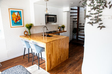 Danish l-shaped dark wood floor and brown floor eat-in kitchen photo in Salt Lake City with a drop-in sink, flat-panel cabinets, white cabinets, wood countertops, white backsplash, stainless steel appliances, an island and brown countertops