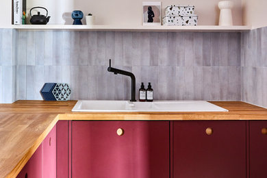 Contemporary l-shaped kitchen in London with a drop-in sink, flat-panel cabinets, red cabinets, wood benchtops, grey splashback, multi-coloured floor and brown benchtop.