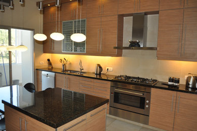 Example of a mid-sized minimalist galley ceramic tile and beige floor enclosed kitchen design in Other with a double-bowl sink, flat-panel cabinets, medium tone wood cabinets, granite countertops, beige backsplash, ceramic backsplash, stainless steel appliances, an island and black countertops