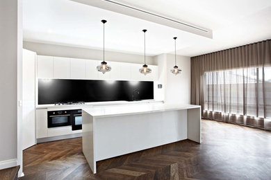 Large contemporary single-wall open plan kitchen in Melbourne with flat-panel cabinets, white cabinets, quartz benchtops, black splashback, black appliances, medium hardwood floors, with island and brown floor.