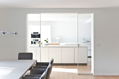 Geschlossene Moderne Küche mit flächenbündigen Schrankfronten, weißen Schränken, Küchenrückwand in Weiß, Glasrückwand, Elektrogeräten mit Frontblende, hellem Holzboden und Kücheninsel in Sonstige