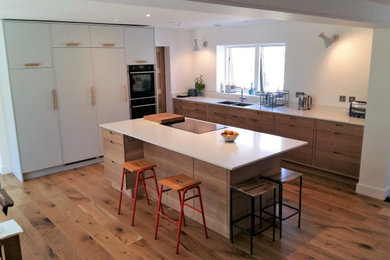 Photo of a large contemporary grey and brown l-shaped open plan kitchen in Cornwall with a double-bowl sink, flat-panel cabinets, medium wood cabinets, composite countertops, black appliances, medium hardwood flooring, a breakfast bar, brown floors and white worktops.