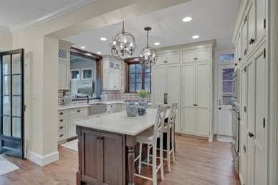 Large french country u-shaped light wood floor and brown floor enclosed kitchen photo in Other with a farmhouse sink, beaded inset cabinets, white cabinets, quartz countertops, brown backsplash, brick backsplash, stainless steel appliances, an island and white countertops