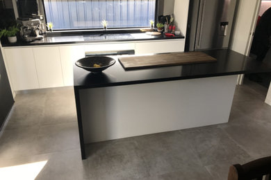 Mid-sized modern galley open plan kitchen in Sydney with a drop-in sink, recessed-panel cabinets, white cabinets, quartz benchtops, black splashback, glass sheet splashback, black appliances, ceramic floors, with island, grey floor and brown benchtop.
