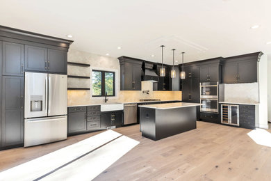 Example of a large transitional l-shaped light wood floor and beige floor open concept kitchen design in Boston with a farmhouse sink, shaker cabinets, black cabinets, granite countertops, beige backsplash, marble backsplash, stainless steel appliances, an island and white countertops