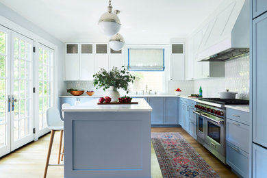 Diseño de cocina clásica renovada con armarios estilo shaker, puertas de armario azules, salpicadero blanco, electrodomésticos de acero inoxidable, suelo de madera en tonos medios, una isla y encimeras blancas