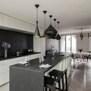 Zweizeilige, Große Moderne Wohnküche mit Unterbauwaschbecken, weißen Schränken, Küchenrückwand in Schwarz, Elektrogeräten mit Frontblende, braunem Holzboden und Kücheninsel in Paris