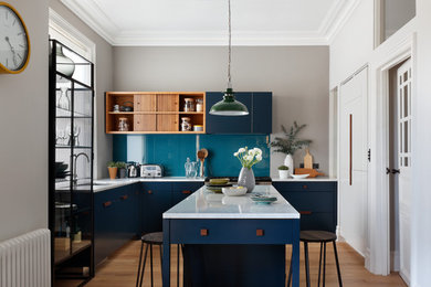 Inspiration for a medium sized contemporary l-shaped kitchen/diner in London with a double-bowl sink, flat-panel cabinets, blue cabinets, marble worktops, blue splashback, glass sheet splashback, stainless steel appliances, light hardwood flooring, an island and white worktops.