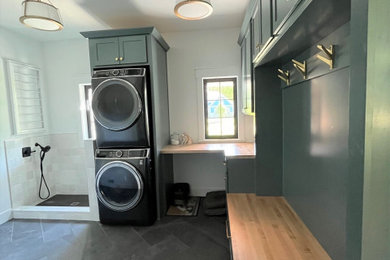 Large transitional l-shaped porcelain tile and gray floor utility room photo in Detroit with shaker cabinets, green cabinets, wood countertops, white backsplash, ceramic backsplash, white walls, a stacked washer/dryer and beige countertops