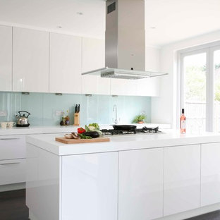 Foto de cocina de galera, minimalista, grande, con armarios con paneles lisos, puertas de armario blancas, encimera de acrílico, salpicadero blanco, salpicadero de vidrio templado, electrodomésticos de acero inoxidable, suelo de madera oscura y una isla