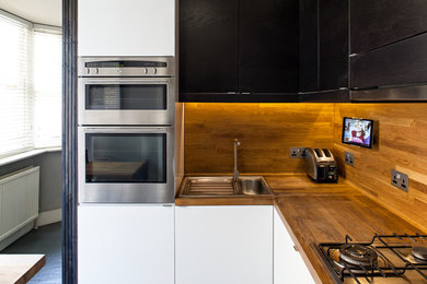 Inspiration for a small transitional l-shaped open plan kitchen in London with a drop-in sink, flat-panel cabinets, black cabinets, wood benchtops, brown splashback, timber splashback, stainless steel appliances, painted wood floors, a peninsula, grey floor and brown benchtop.