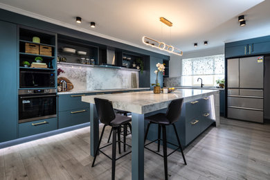 This is an example of a large contemporary l-shaped eat-in kitchen in Melbourne with a drop-in sink, open cabinets, blue cabinets, quartz benchtops, white splashback, stone slab splashback, black appliances, with island, grey floor and white benchtop.