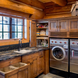 Modelo de cuarto de lavado en L, rústico, grande, con fregadero bajoencimera, armarios con paneles empotrados, puertas de armario de madera oscura, encimera de granito, suelo de pizarra y lavadora y secadora juntas