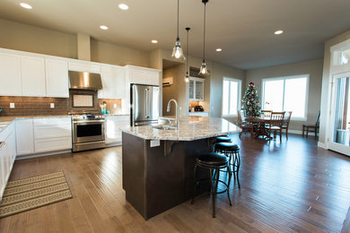 Large transitional l-shaped medium tone wood floor and brown floor open concept kitchen photo in Seattle with an undermount sink, shaker cabinets, white cabinets, granite countertops, brown backsplash, subway tile backsplash, stainless steel appliances and an island