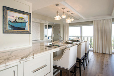 Design ideas for a large beach style galley open plan kitchen in Orlando with a double-bowl sink, glass-front cabinets, white cabinets, marble benchtops, white splashback, stone slab splashback, stainless steel appliances and light hardwood floors.