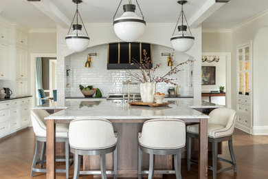 Transitional u-shaped medium tone wood floor, brown floor and exposed beam kitchen photo in New York with a farmhouse sink, shaker cabinets, white cabinets, white backsplash, subway tile backsplash, stainless steel appliances, an island, marble countertops and gray countertops