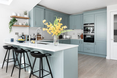 Design ideas for a traditional u-shaped kitchen/diner in London with a submerged sink, shaker cabinets, blue cabinets, engineered stone countertops, white splashback, engineered quartz splashback, integrated appliances, light hardwood flooring, a breakfast bar, beige floors and white worktops.