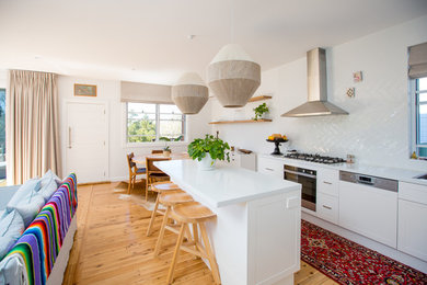 This is an example of a beach style l-shaped open plan kitchen in Other with an undermount sink, shaker cabinets, white cabinets, white splashback, stainless steel appliances, medium hardwood floors, with island, brown floor and white benchtop.