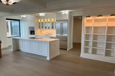 Transitional medium tone wood floor, brown floor and coffered ceiling kitchen photo in New York with a drop-in sink, raised-panel cabinets, white cabinets, marble countertops, white backsplash, marble backsplash, stainless steel appliances, an island and white countertops