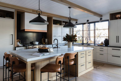 Photo of a large coastal l-shaped open plan kitchen in Portland with a submerged sink, recessed-panel cabinets, white cabinets, granite worktops, black splashback, granite splashback, black appliances, light hardwood flooring, an island, black worktops, exposed beams and brown floors.