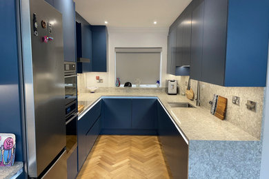Medium sized contemporary u-shaped open plan kitchen in Essex with a single-bowl sink, flat-panel cabinets, blue cabinets, quartz worktops, grey splashback, engineered quartz splashback, light hardwood flooring, no island, brown floors, grey worktops and feature lighting.