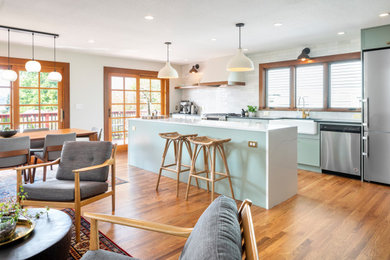 Mid-sized trendy single-wall medium tone wood floor open concept kitchen photo in Seattle with a farmhouse sink, shaker cabinets, green cabinets, quartz countertops, white backsplash, ceramic backsplash, stainless steel appliances, an island and white countertops
