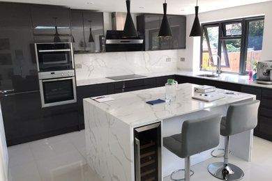 Large modern l-shaped open plan kitchen in London with a drop-in sink, flat-panel cabinets, black cabinets, quartzite benchtops, white splashback, black appliances, carpet, with island and white benchtop.