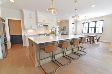 Example of a large transitional l-shaped light wood floor and beige floor open concept kitchen design in DC Metro with a farmhouse sink, shaker cabinets, white cabinets, quartz countertops, white backsplash, ceramic backsplash, stainless steel appliances, an island and white countertops