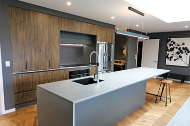Inspiration for a contemporary galley kitchen in Christchurch with an undermount sink, brown cabinets, solid surface benchtops, black splashback, stone tile splashback, stainless steel appliances, laminate floors, with island, brown floor and grey benchtop.