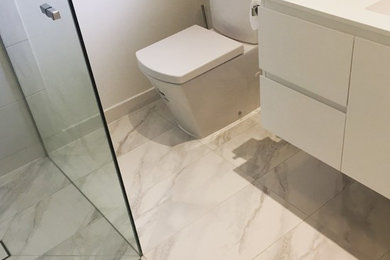 Photo of a small modern bathroom in Melbourne with a corner shower, a one-piece toilet, white tile, ceramic tile, white walls, porcelain floors, an integrated sink, white floor, a hinged shower door, flat-panel cabinets, white cabinets and marble benchtops.