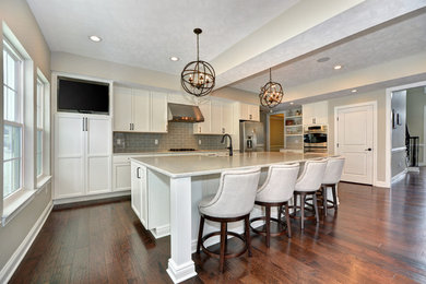 Example of a mid-sized classic single-wall dark wood floor and brown floor eat-in kitchen design in Other with a farmhouse sink, recessed-panel cabinets, white cabinets, granite countertops, gray backsplash, subway tile backsplash, stainless steel appliances, an island and white countertops