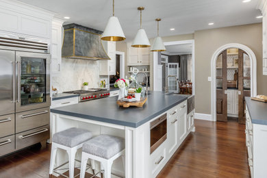 Example of a transitional galley medium tone wood floor and brown floor kitchen design in Philadelphia with a farmhouse sink, shaker cabinets, white cabinets, white backsplash, stone slab backsplash, stainless steel appliances, an island and gray countertops