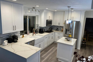 Example of a mid-sized minimalist u-shaped laminate floor and gray floor kitchen pantry design with shaker cabinets, white cabinets, quartz countertops, an island, white countertops, a farmhouse sink, blue backsplash, glass tile backsplash and stainless steel appliances