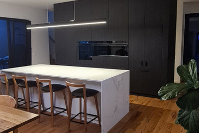 This is an example of a large contemporary galley open plan kitchen in Geelong with a double-bowl sink, dark wood cabinets, solid surface benchtops, white splashback, ceramic splashback, black appliances, medium hardwood floors, with island and white benchtop.
