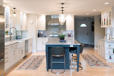 Transitional u-shaped light wood floor open concept kitchen photo in New York with a farmhouse sink, white cabinets, quartzite countertops, gray backsplash, ceramic backsplash, stainless steel appliances, an island, gray countertops and shaker cabinets