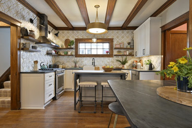 Inspiration for a transitional u-shaped medium tone wood floor, brown floor and exposed beam eat-in kitchen remodel in Dallas with a farmhouse sink, shaker cabinets, white cabinets, metallic backsplash, stainless steel appliances, an island and black countertops